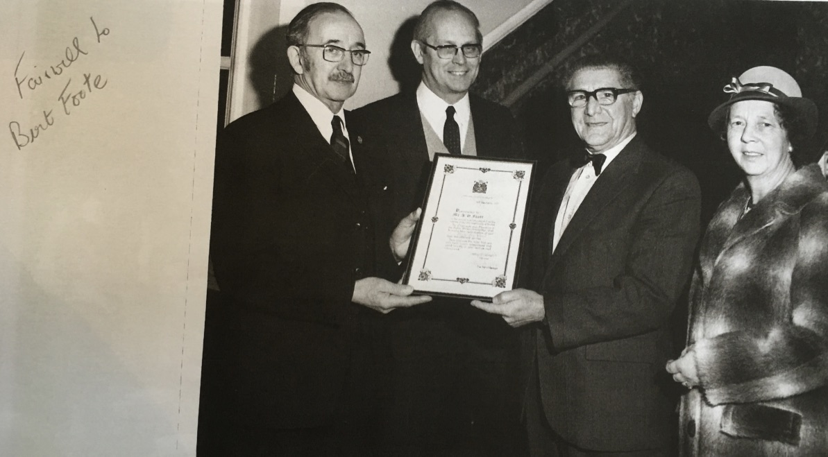 Photo of smartly dressed gentlemen and woman, as a framed certificate is passed