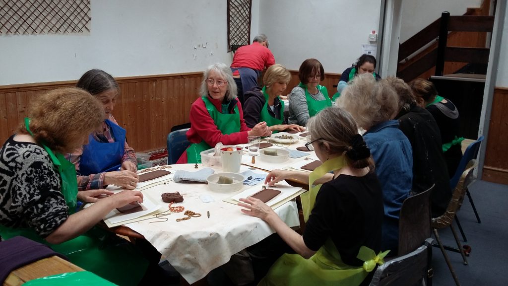 10 people around a table, wearing colourful plastic aprons, each working withclay