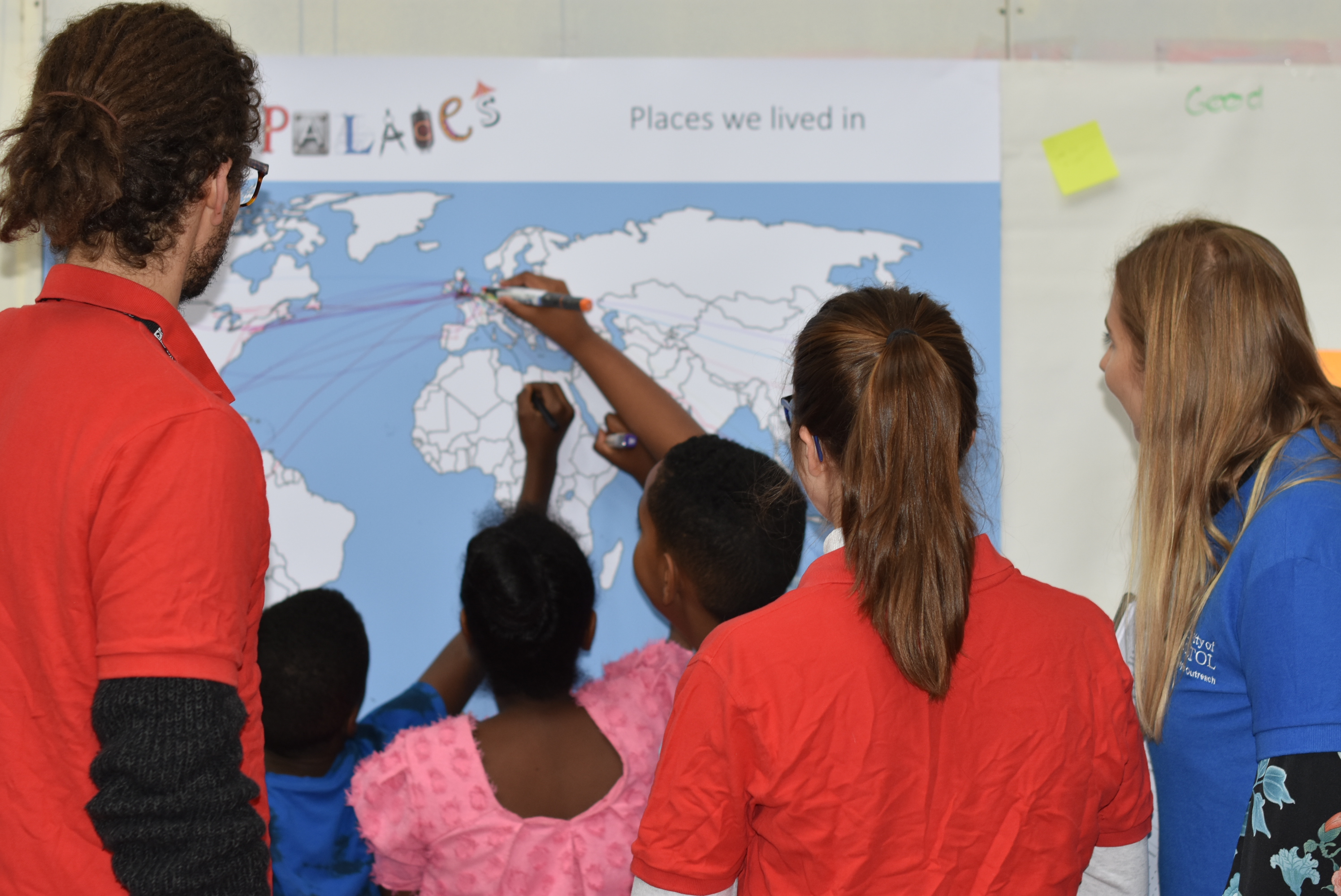 Children drawing lines from across a world map to the UK, watched by adults inbright t-shirts