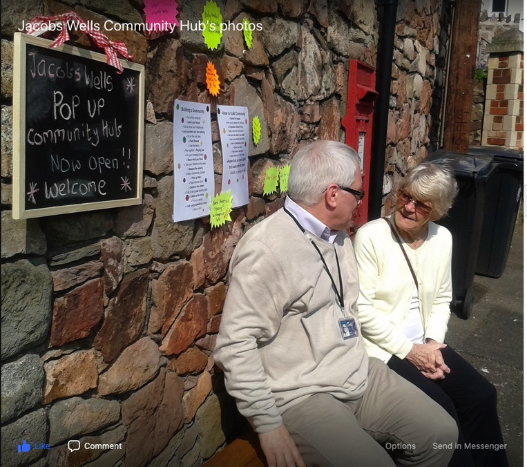 Two people sitting by a stone wall. Attached to the wall are blackboard saying “Jacobs Wellspopup community hub now open!! Welcome,” and other paper notices about the hub